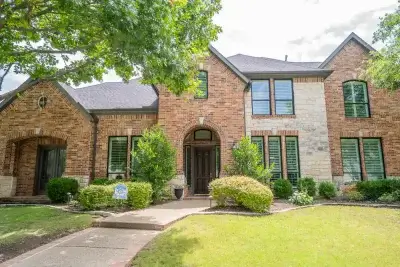 Two-story brick and stone house with black shutters, surrounded by green trees, bushes, and a well-kept lawn.