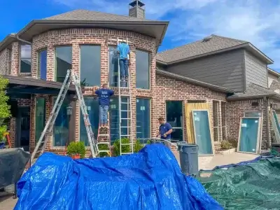 Three workers replacing windows on a brick house, with ladders and tarps covering the ground outside.
