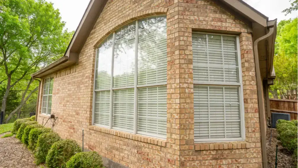 Brick house with large arched windows, white blinds, and bushes lining the exterior; trees in the background.