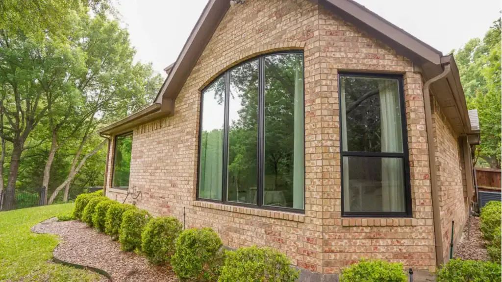 A brick house with large black-trimmed windows, surrounded by green bushes and trees.