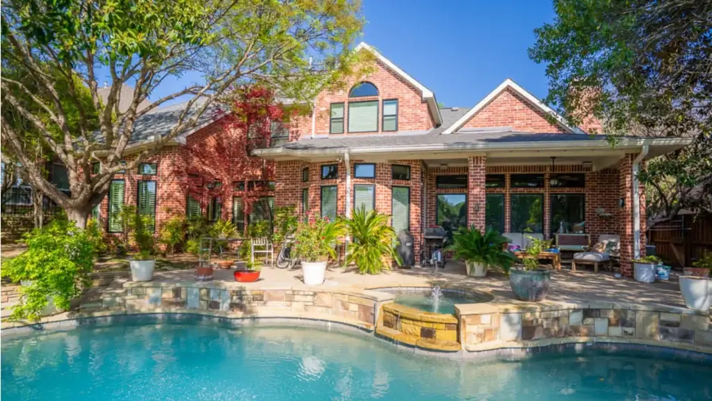 Large brick house with arched windows, patio, potted plants, and a swimming pool in the backyard on a sunny day.