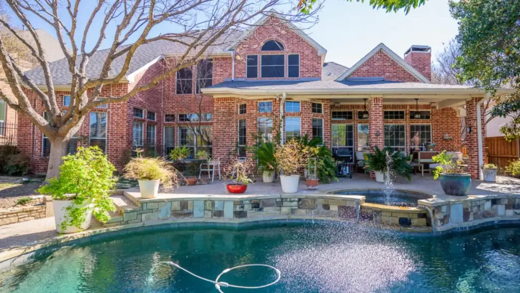 Backyard view of a brick house with large windows, covered patio, potted plants, and a pool with a fountain.