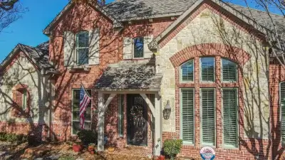Two-story brick and stone house with large windows, an American flag, and trees casting shadows on the facade.