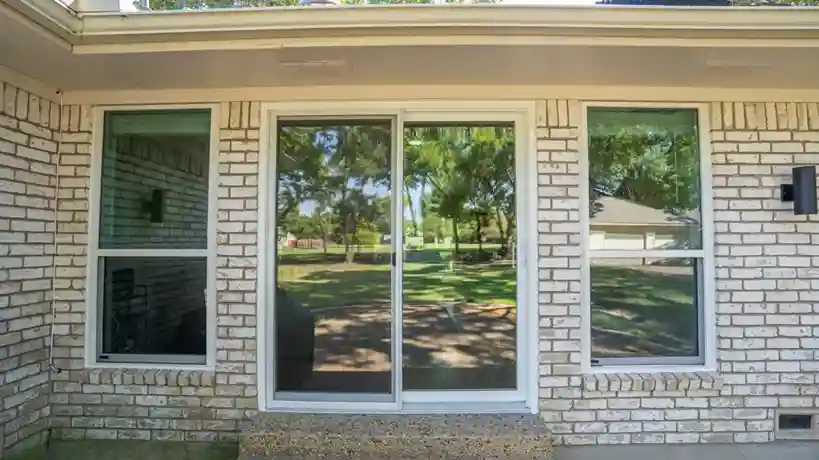 White brick house with two windows and a central glass sliding door reflecting a green yard and trees.