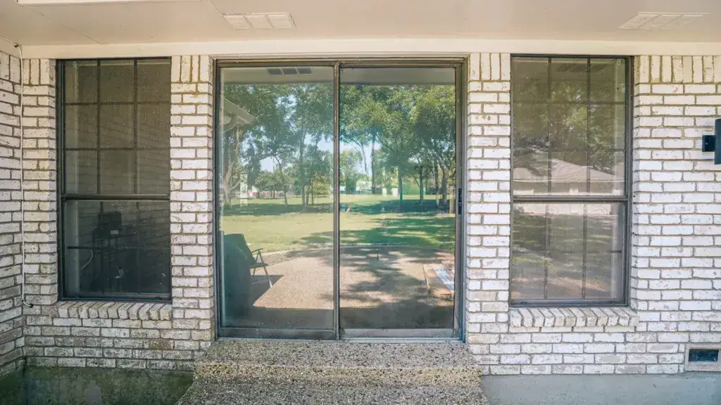 Sliding glass door with two large windows on either side, set in a white brick wall, overlooking a grassy yard.