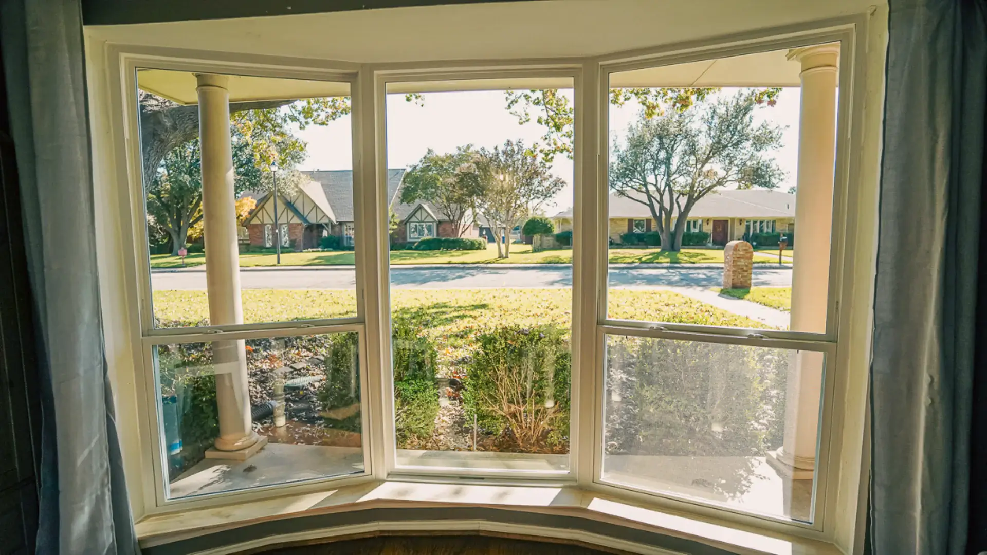 View through a bay window showing a sunny suburban street with trees, grass, and houses across the road.
