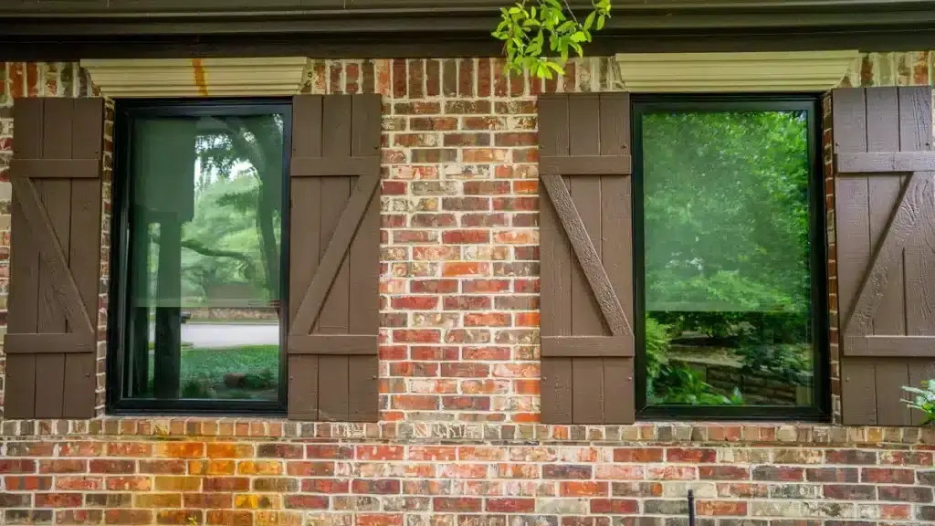 Two windows with brown wooden shutters on a red brick wall, reflecting trees and greenery.