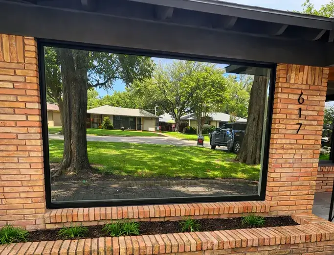 A large, reflective window on a brick house mirrors trees, grass, and homes across a suburban street.
