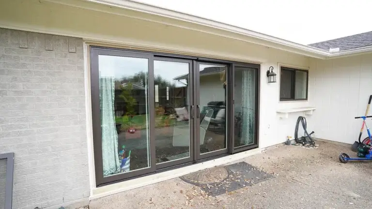 Glass sliding patio door with black frame, reflecting outdoor scene, next to a white wall and concrete patio.