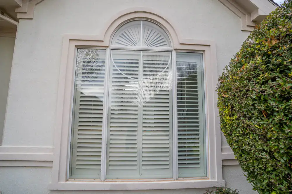 White arched window with shutters on a light-colored house, next to a green, trimmed bush.