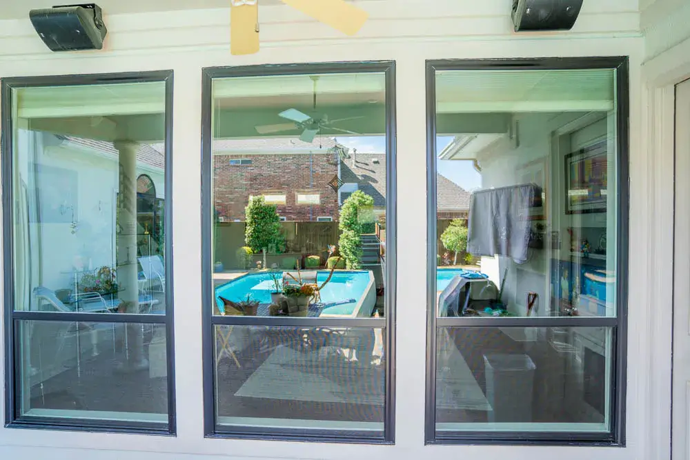 Three large windows reflect and reveal a backyard pool, patio, and greenery on a sunny day.