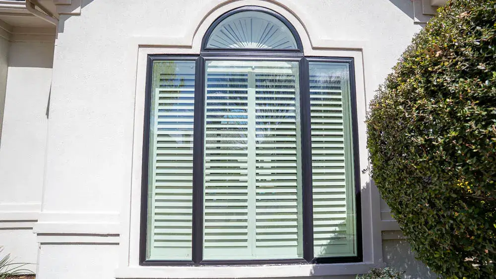 Large arched window with white shutters, set in a light-colored wall, next to a green bush.