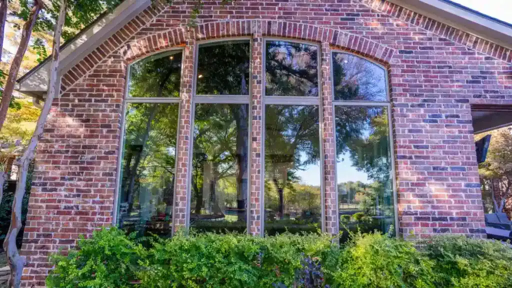Large three-pane arched windows on a brick house, reflecting trees, with green shrubs below.
