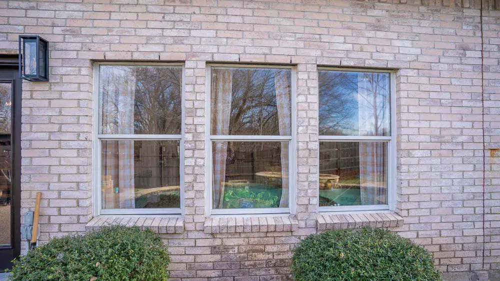 Three white-framed windows on a light brick wall, with bushes in front and trees reflected in the glass.