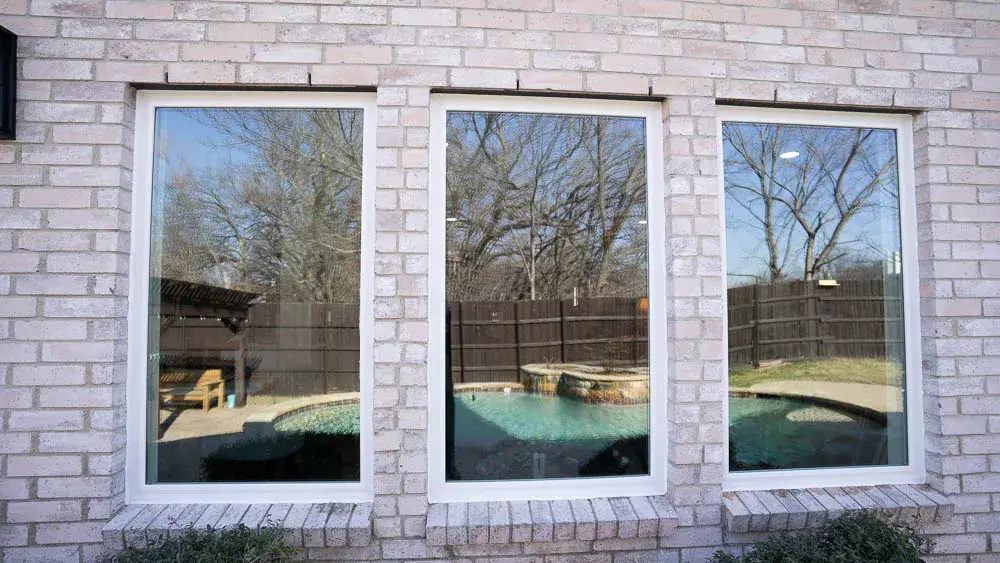 Three large windows in a brick wall reflect a backyard pool, trees, and a wooden fence under a clear sky.