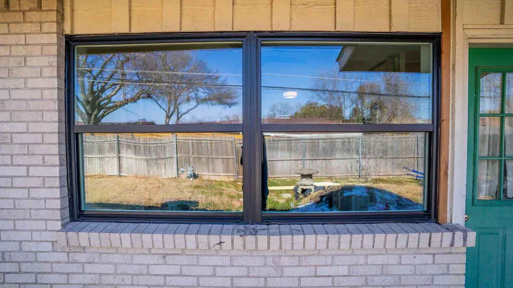 A double-pane window reflects a backyard with a wooden fence, trees, grass, and a garden pond.