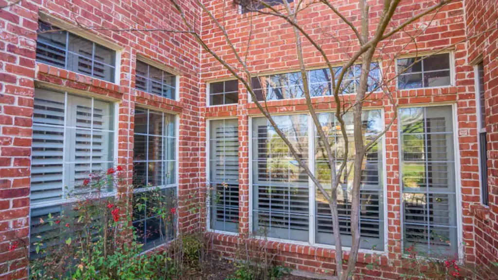 Red brick house with large windows and white shutters, small garden, and bare tree branches in front.