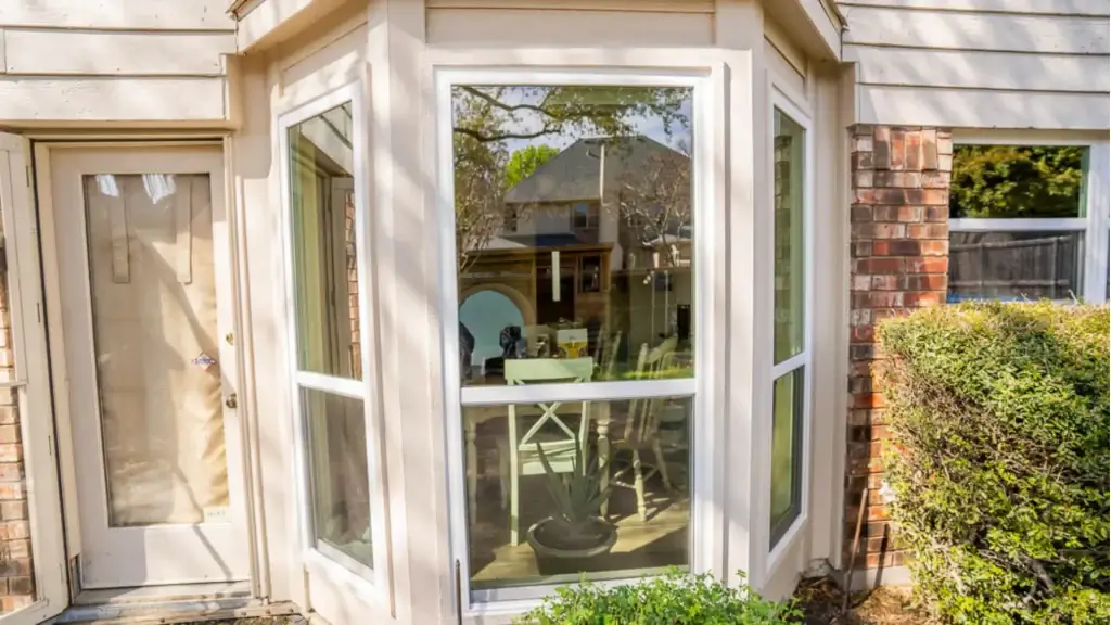 Bay window with white trim, reflecting a patio view; plants and a table are visible inside, bush is next to window.