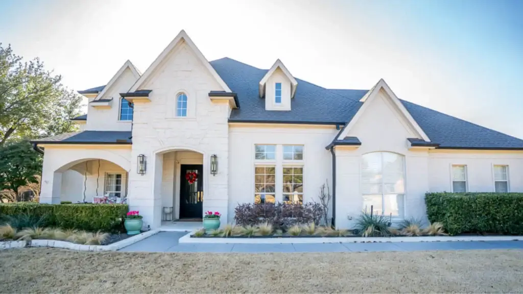 Two-story white house with gabled roof, front porch, large windows, and shrubs along the walkway.
