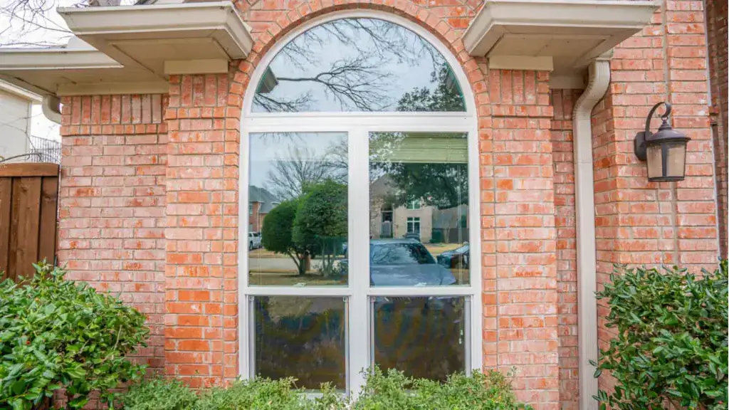 A large arched window set in a red brick wall, with bushes below and a black lamp on the right side.