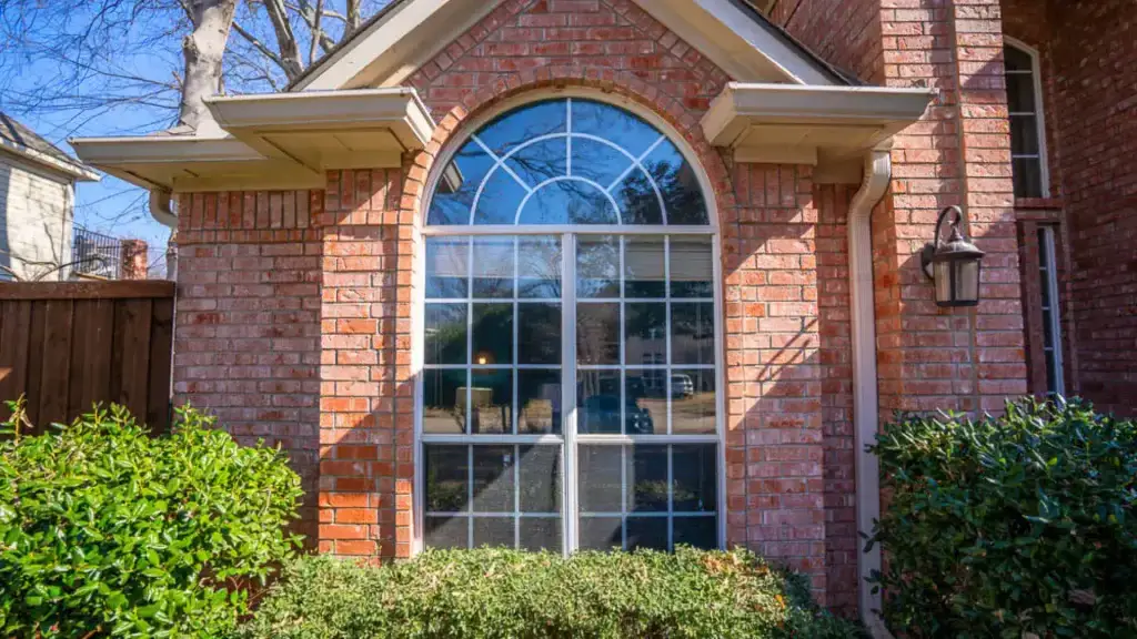Large arched window with grid panes on a red brick house, surrounded by bushes and greenery.
