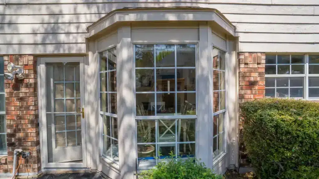 Bay window and glass door on a brick house exterior with bushes in front and sunlight casting shadows.