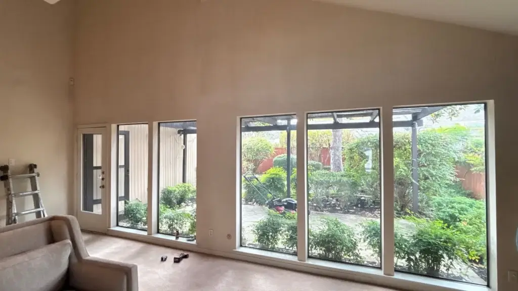 Five large windows and a door overlook a patio garden from a living room with beige walls, a sofa, and a ladder.