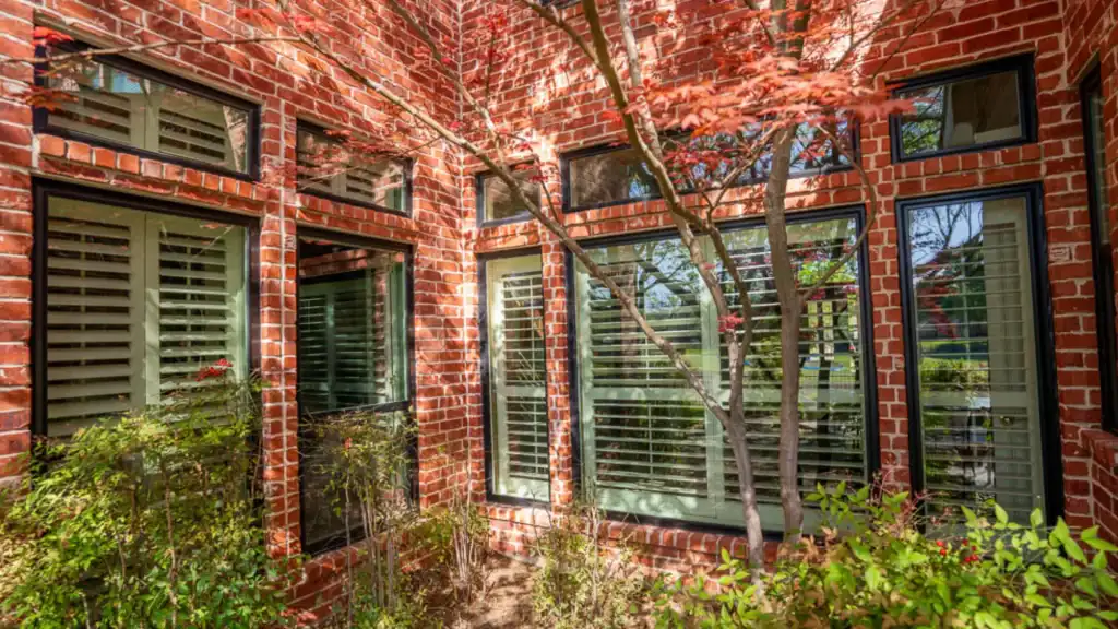 Red brick house exterior with large windows, white shutters, and leafy trees casting shadows in a garden area.