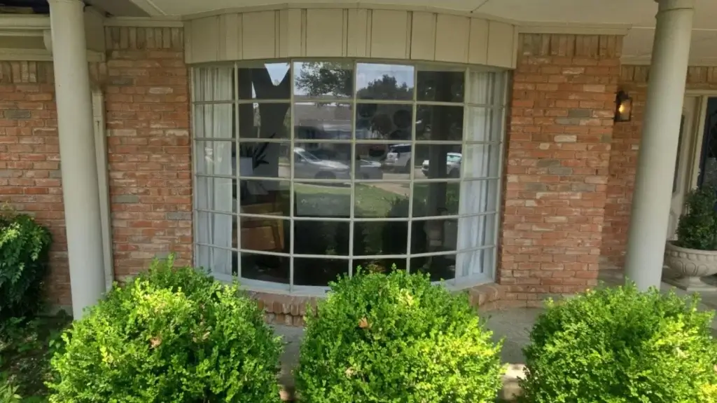 A large bay window with grid panes on a brick house, with green bushes in front and white curtains inside.