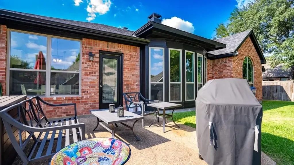 Brick patio with outdoor seating, a covered grill, large windows, and a colorful bowl in a backyard setting.