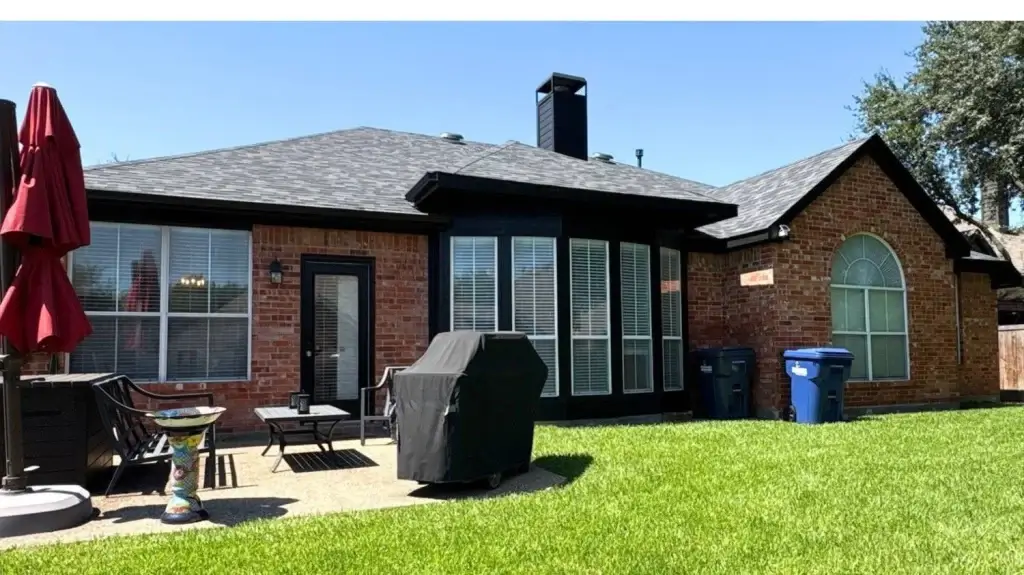 A brick house with a patio, barbecue grill, patio furniture, red umbrella, and green lawn on a sunny day.