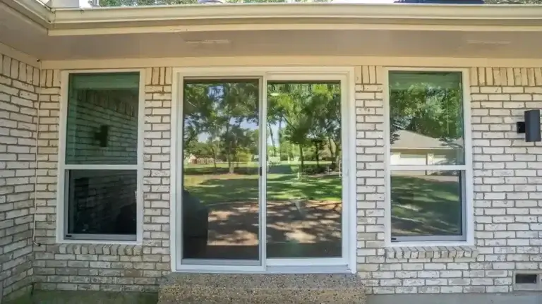 Sliding glass door and two windows in a white brick wall reflect trees and a grassy yard outside.