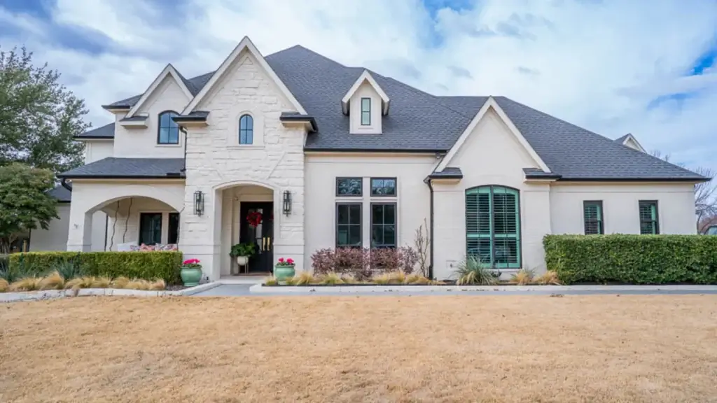 Modern two-story house with light stone exterior, dark roof, large windows, and a landscaped front yard.