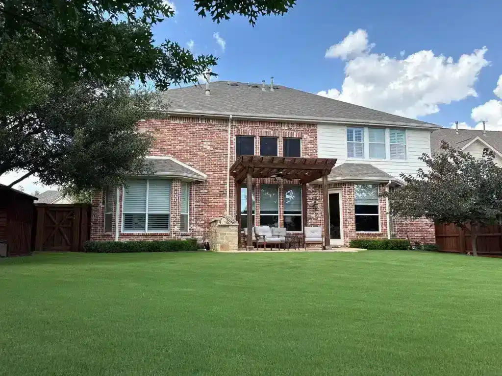 Red-brick two-story house with pergola, patio furniture, and green lawn under a partly cloudy sky.