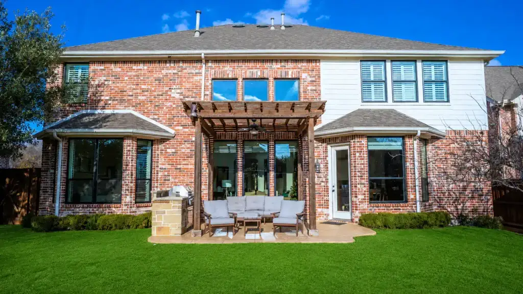Two-story brick house with pergola-covered patio, outdoor seating, and well-manicured green lawn on a sunny day.