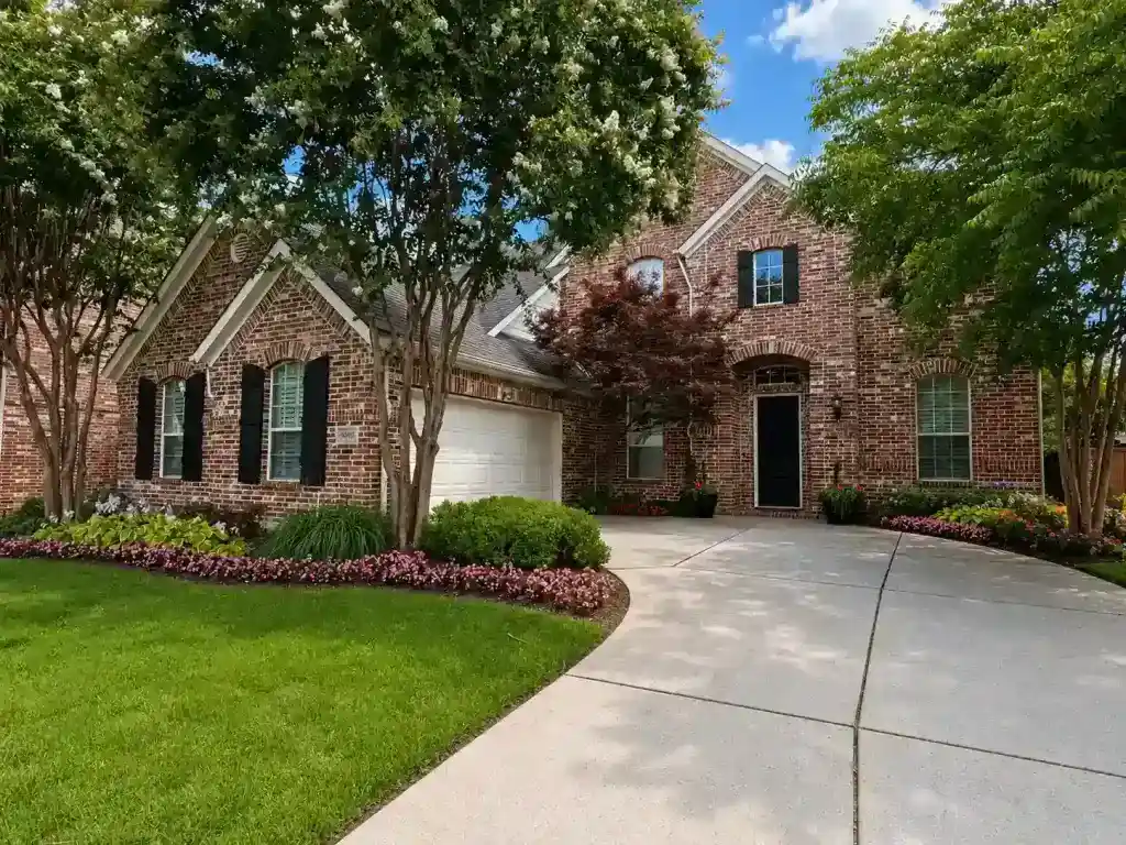 Brick house with white garage, green lawn, trees, shrubs, and a curved driveway under a partly cloudy blue sky.