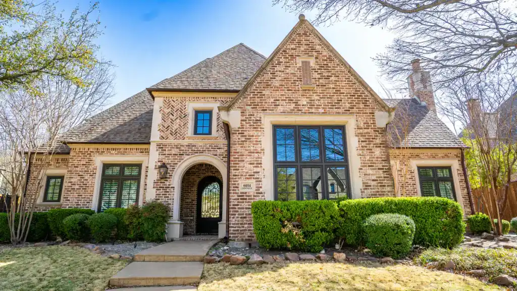 Brick house with tall gabled roof, arched entryway, and manicured bushes in front yard under a clear blue sky.