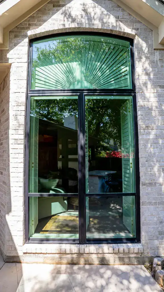 Large arched window with sunburst pattern, reflecting trees and sky, set in a light brick exterior wall.