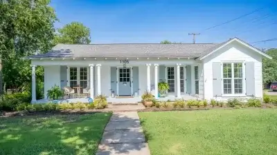White single-story house with a front porch, columns, plants, and a green lawn under a clear blue sky.