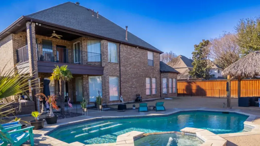 Two-story brick house with a balcony, pool, hot tub, patio seating, and a thatched-roof cabana in the backyard.