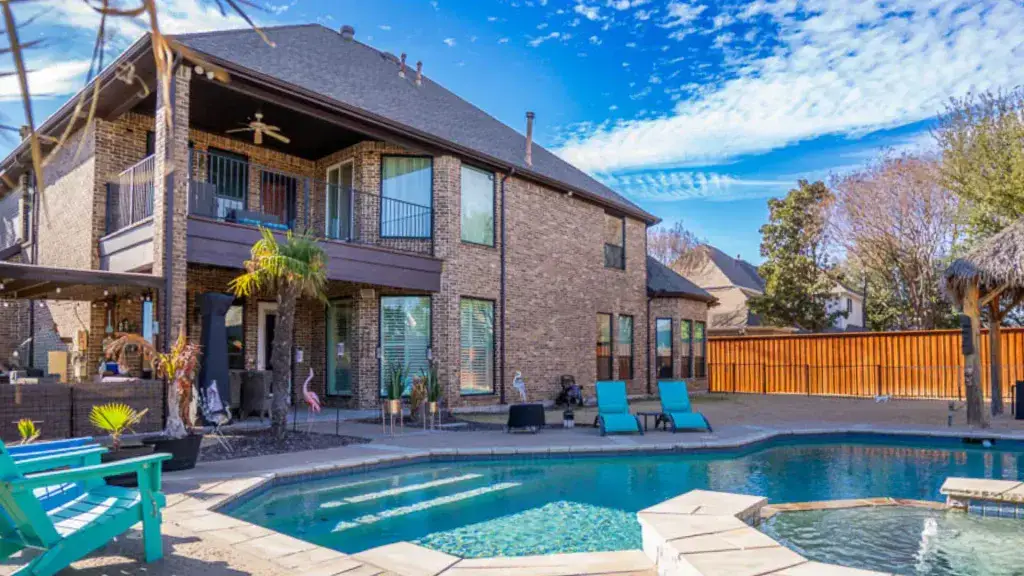 Two-story brick house with backyard pool, patio seating, palm trees, and a wooden fence under a blue sky.