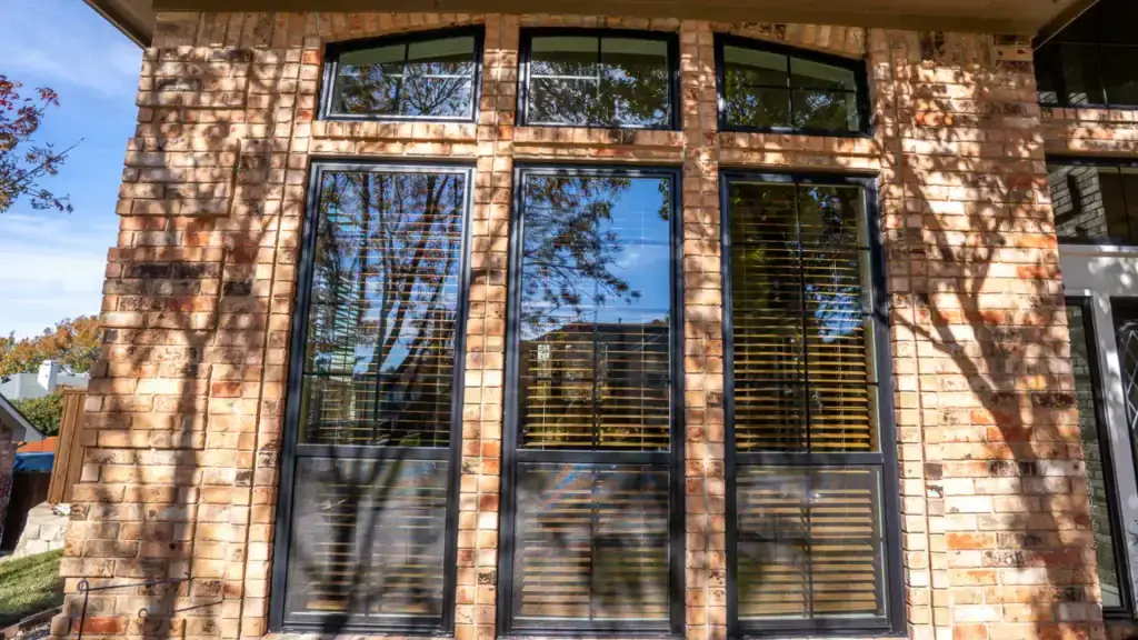 Tall windows with black frames reflect trees and a house, set in a brick wall with sunlight and shadows visible.