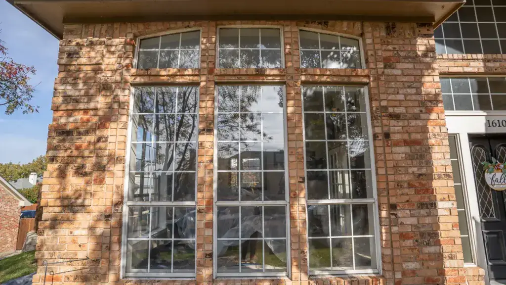 Large brick house window with four tall window panels reflecting trees and sky, with white curtains inside.