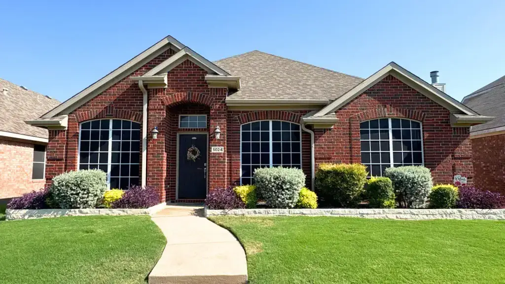 Single-story red brick house with large windows, a dark front door, and neatly trimmed bushes and green lawn.