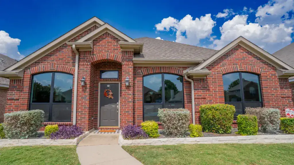 Single-story red brick house with arched windows, shrubs, and a front path under a bright blue sky with clouds.