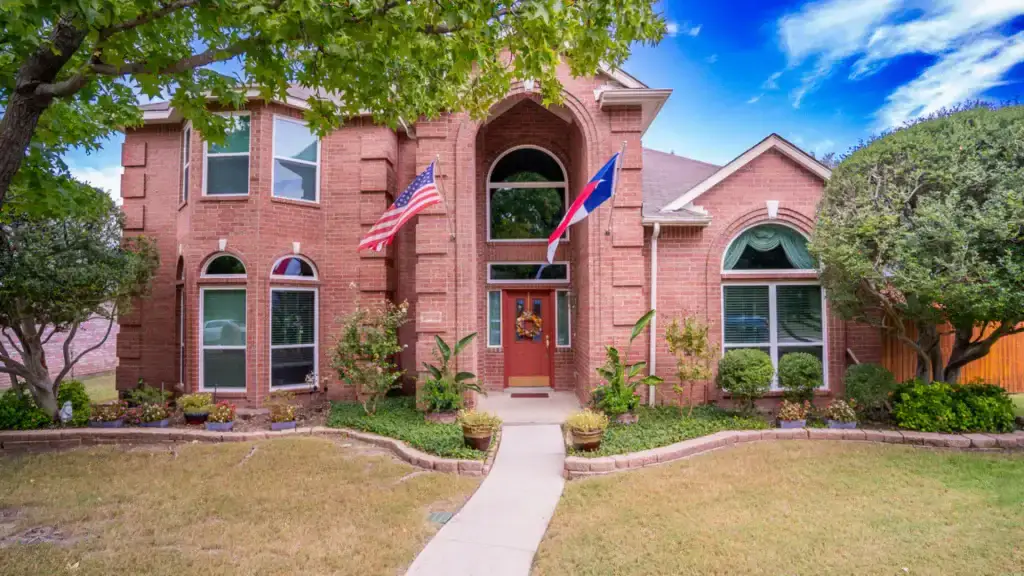 Two-story red brick house with American and Texas flags, lush greenery, and clear blue sky overhead.