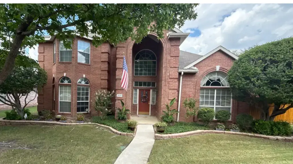 Two-story red brick house with arched windows, an American flag, and a well-kept front yard with trees and shrubs.