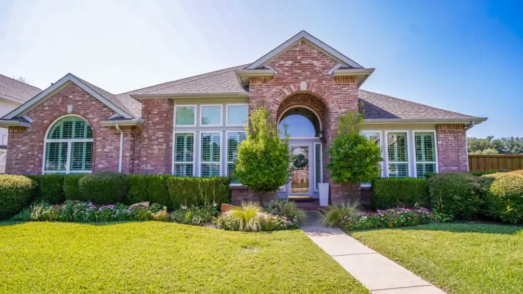 Single-story brick house with arched entry, large windows, green lawn, and trimmed bushes under a clear blue sky.