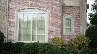 Red brick house with two windows featuring white trim and shutters, surrounded by green bushes and shrubs.
