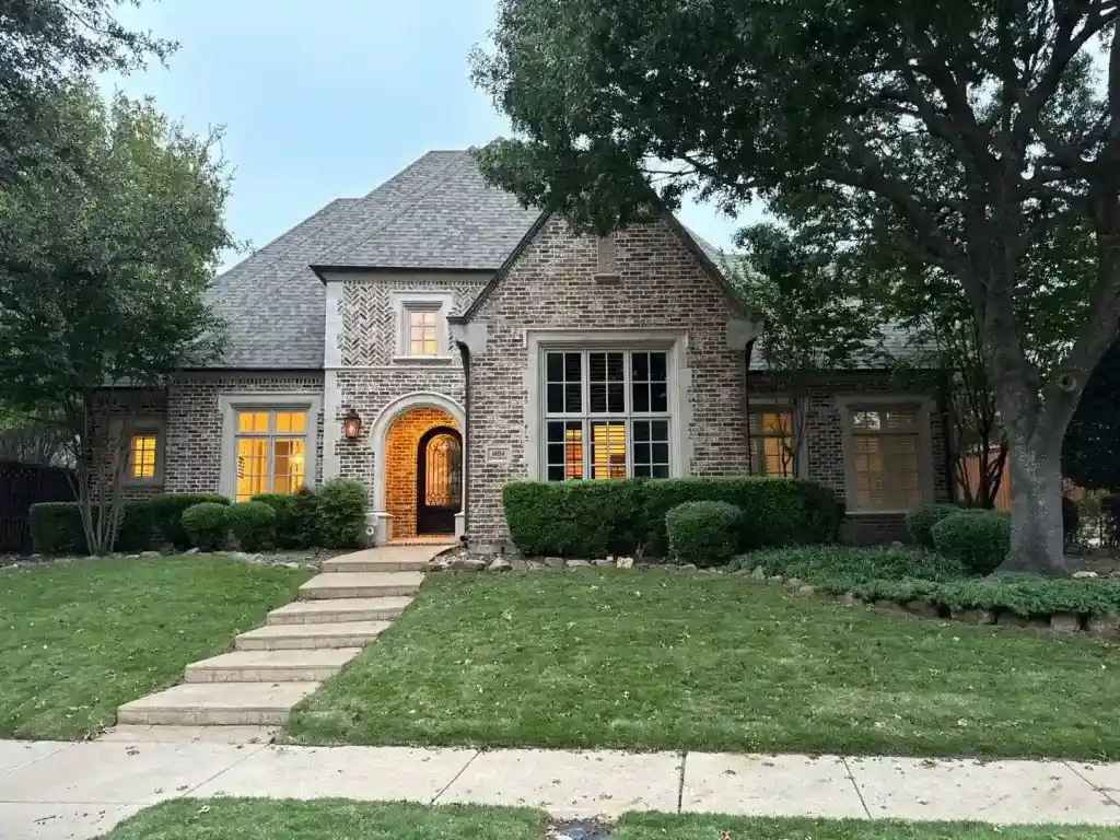 A brick house with large windows, lit inside, surrounded by trees and a neatly trimmed lawn at dusk.